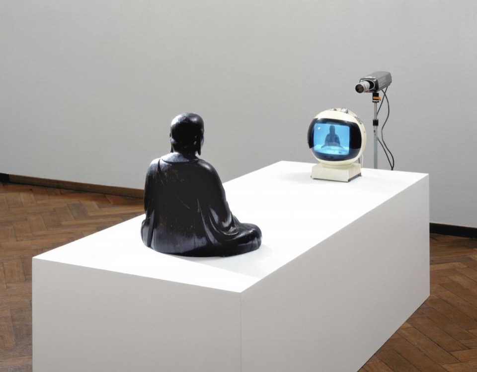 A seated Buddha faces a single television showing its live image, with a camera behind the screen.